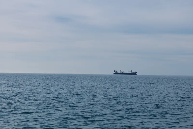 A lone cargo ship sails across a calm sea toward the distant horizon, highlighting vast ocean expanse, maritime travel, and tranquil open water under a soft sky.