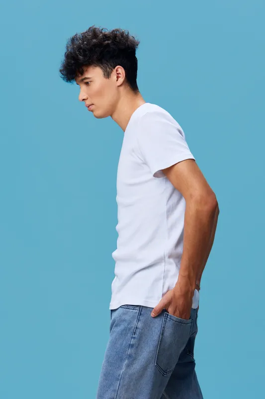 young man with curly hair wearing white t-shirt and blue jeans standing sideways against solid blue background in casual style