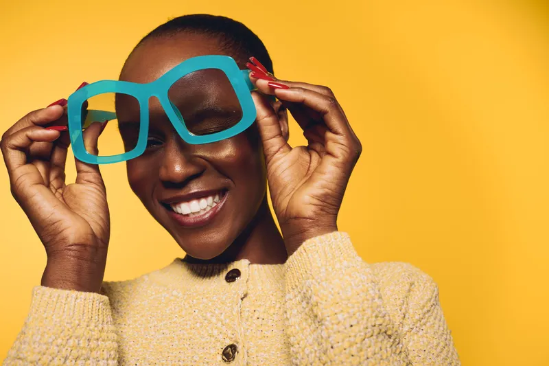Happy black woman with dark skin smiling and holding large blue paper glasses on yellow background. Concept of joy, fashion, and playful portrait of African ethnicity female model.
