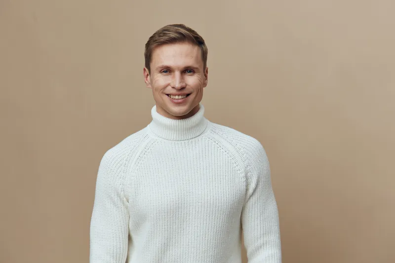 Smiling man wearing white knitted sweater looking at camera isolated on plain beige background studio portrait. People lifestyle concept, happiness, casual fashion, warmth, cheerful expression, male m