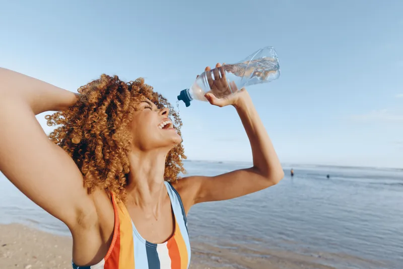 Young woman with curly hair on the beach drinks water from a bottle during a bright sunlit moment, wearing a striped swimsuit and enjoying a relaxed, carefree vibe