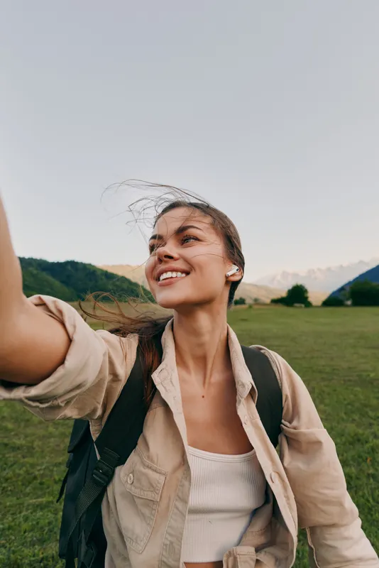 Young woman with long hair smiles happily while taking a selfie outdoors in a green field with mountains in the background on a clear day.