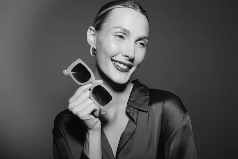 Smiling Caucasian woman with light skin holding sunglasses wearing satin shirt. Studio portrait with elegant and joyful expression.