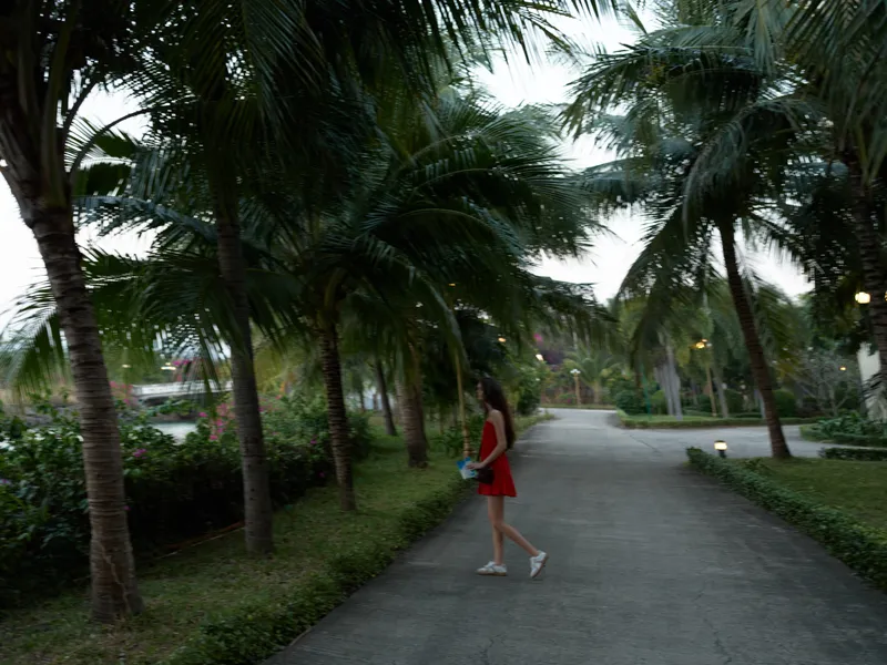 Woman in red dress walking along tropical pathway surrounded by palm trees during dusk, peaceful evening with greenery and soft natural light.