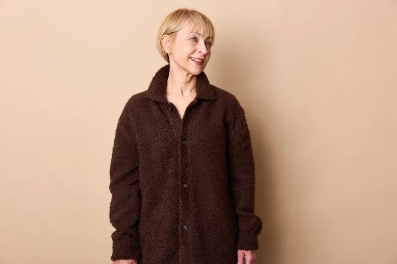 Elderly woman smiling, studio portrait, wearing brown coat, standing against beige background, fashion casual look, warm calm expression, minimal indoor wall, light, copy space
