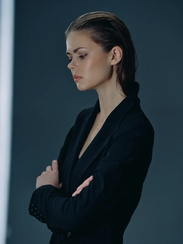 Serious woman in black jacket posing with arms crossed against dark background.