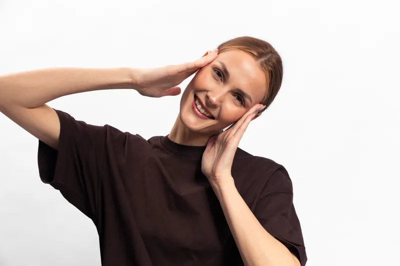 Smiling young woman with brown hair wearing a dark t shirt poses with hands framing her face on white background. Concept of happiness, joy, and positive emotions in casual style.