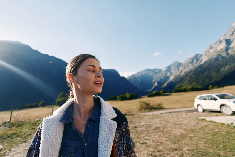 Woman, portrait, mountains, car, nature, travel, outdoors. Young woman in a cozy coat breathes in sunlight with eyes closed at a roadside mountain field next to parked car during scenic road trip.