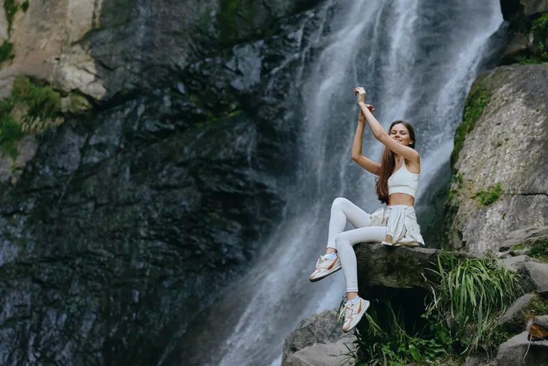Young woman hiking near a waterfall, wearing white activewear and sneakers, seated on a rock, enjoying nature with lush greenery and flowing water in the background.