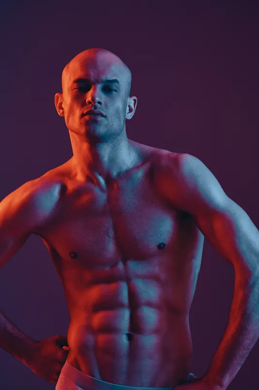 Athletic man posing confidently with strong physique, illuminated by dynamic blue and red light, showcasing strength and power in a minimalistic background.