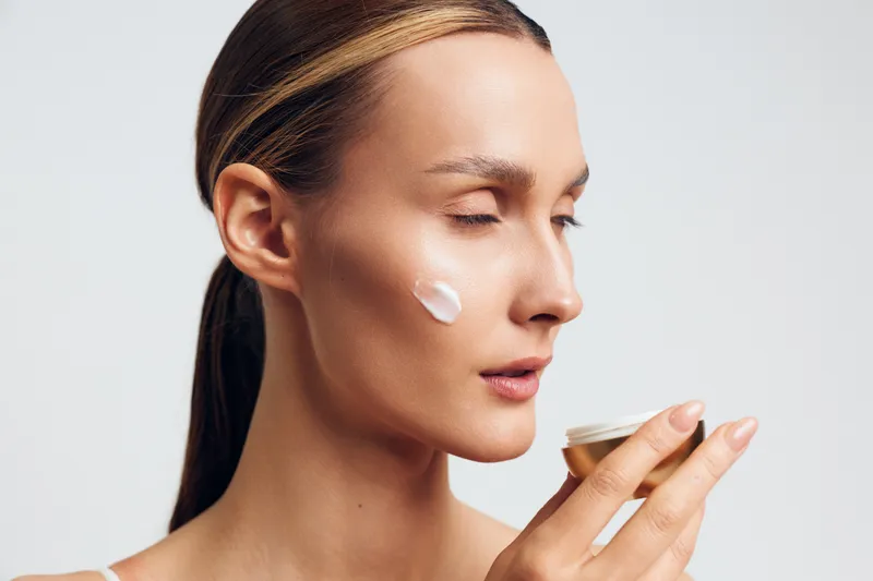 A young woman with clear skin applies white cream on her cheek while holding a small jar. The close-up shows skincare routine and natural beauty concept with soft lighting.