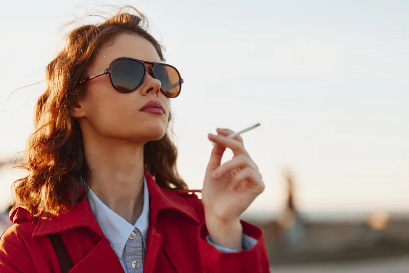 A stylish woman in a red coat wears sunglasses, smoking outdoors against a bright sky. A bold urban portrait conveying confidence, attitude, and street fashion.