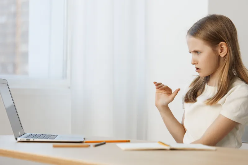 Young girl engaging in an online class with a serious expression, wearing a white shirt, surrounded by a bright, minimalistic background, reflecting focus and dedication