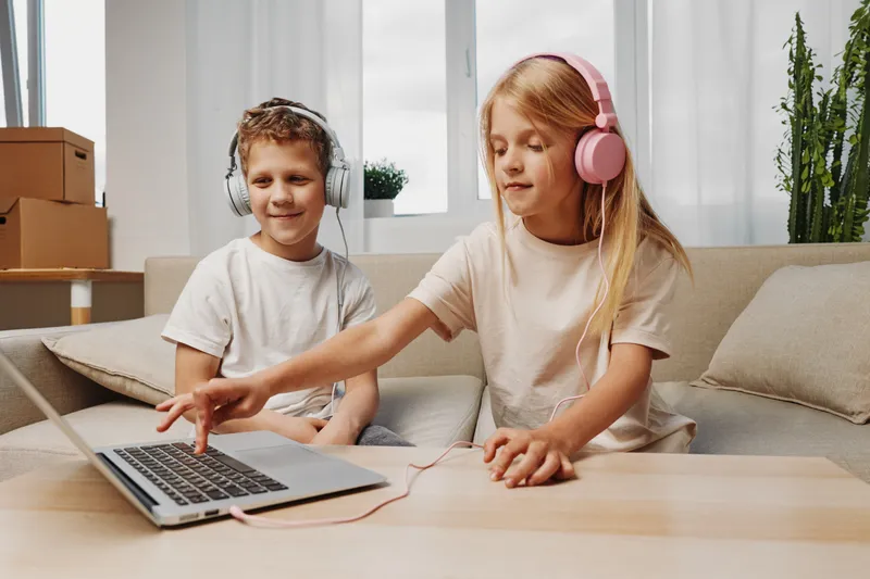 Two children wearing headphones listen to music on a laptop in a cozy living room with natural light. Concept of childhood, technology, entertainment, and home leisure activities.