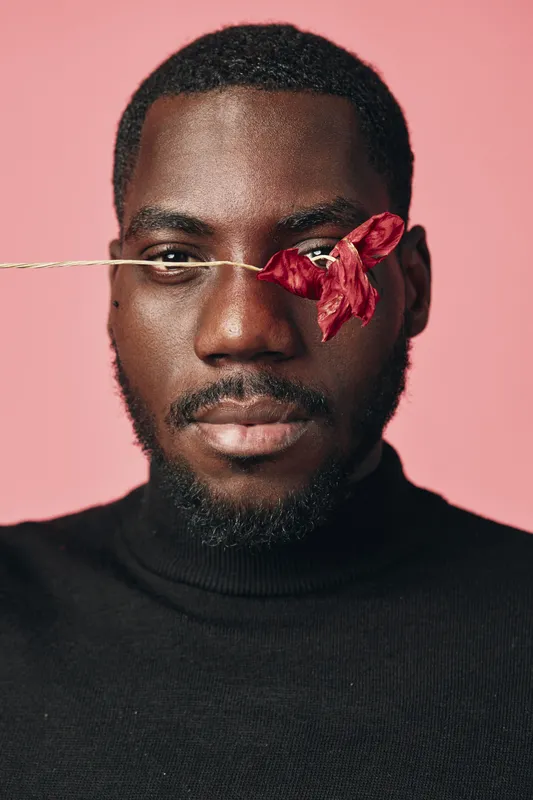 Black man with beard and short hair wearing black turtleneck with dried red flower covering one eye on pink background in creative conceptual visual style portrait.
