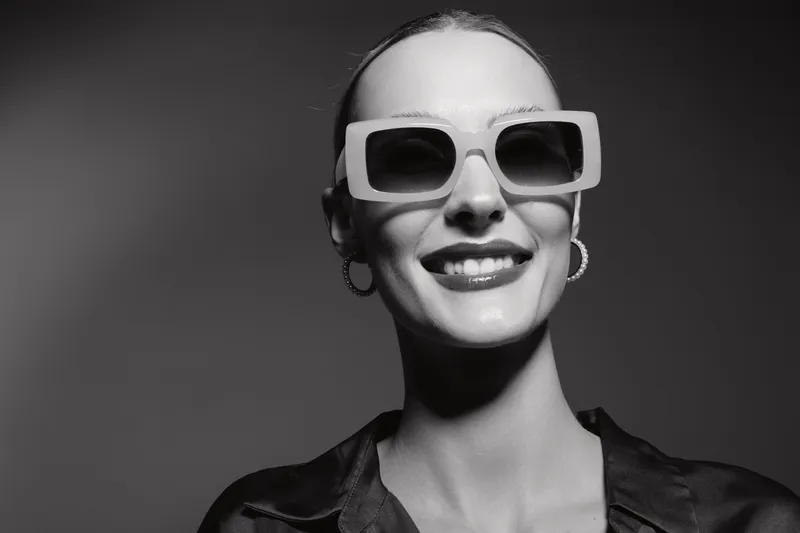 Smiling Caucasian woman with fair skin wearing large square sunglasses and hoop earrings in black and white studio portrait. Concept of fashion, style, and confidence.