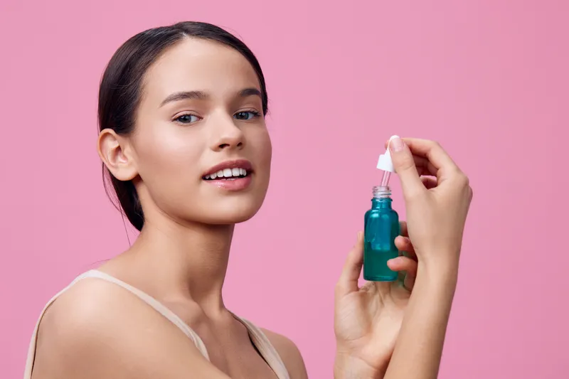Young woman applying skincare serum against a pink background, emphasizing beauty and self care with a focus on skin health and radiance