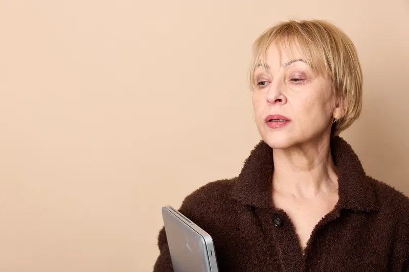 Senior woman blonde portrait indoors beige background dark coat holding notebook calm thoughtful expression soft studio light copy space gentle closeup neutral pose