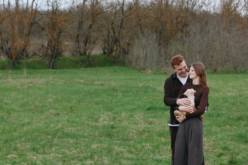 A young couple embraces outdoors while the woman holds a small baby goat in her arms on a green field with bare trees in the background during early spring.