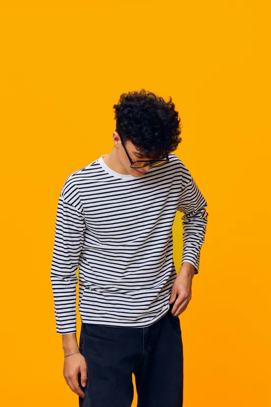 young man wearing glasses and striped shirt looking down against bright yellow background, casual style, curly hair, thoughtful pose, modern fashion