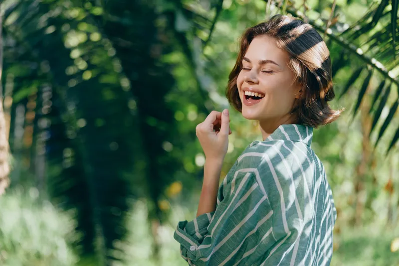Joyful portrait of a woman outdoors, turning with a bright smile, sunlit garden setting and relaxed striped shirt in a candid moment.