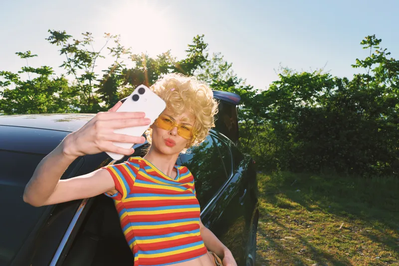 Cheerful woman outdoors, laughing softly, wearing a striped top, taking a selfie beside a car, sunlit greenery in the background, relaxed pose, vintage color mood, fresh vibe