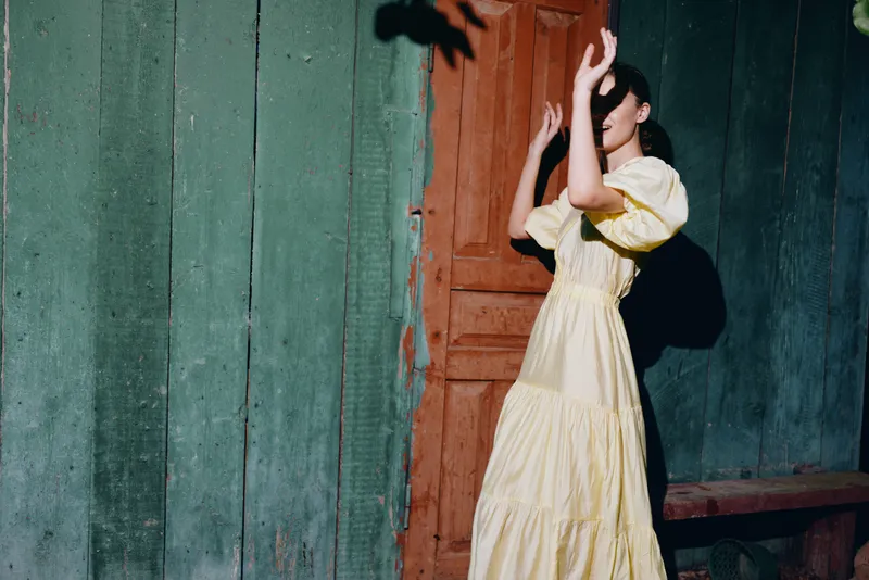 A woman in a pale yellow dress stands beside a green wall and a wooden door, posing with her hands raised, sunlight casting a bold shadow in a vintage outdoor scene.