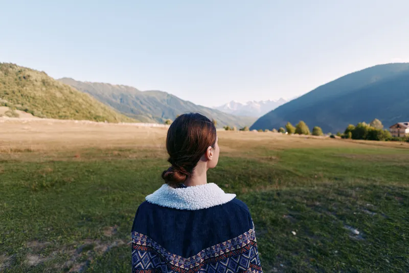 Woman back in sweater stands in wide valley landscape facing distant mountains, nature travel scene with serene meadow and alpine ridges, outdoor exploration and peaceful solitude.