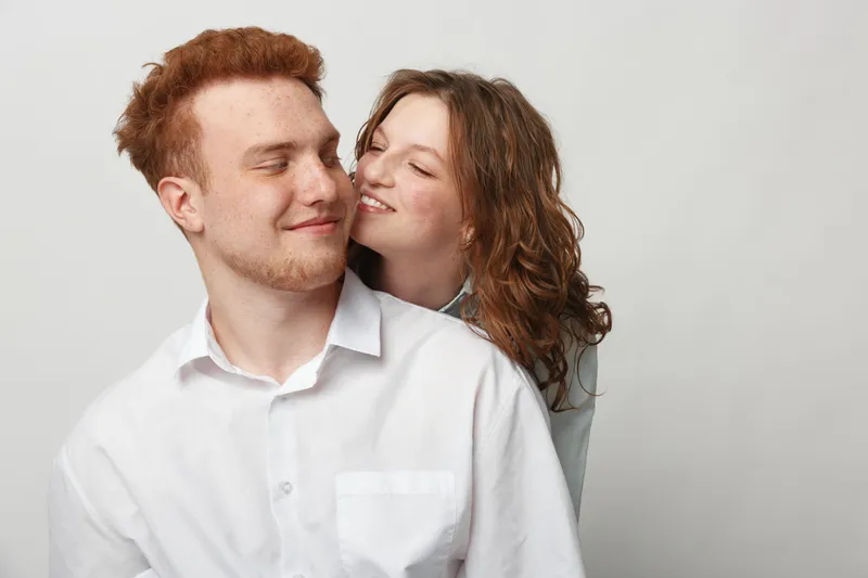 Happy young couple with red hair and curly hair smiling and embracing each other on a light background. Concept of love, affection, relationship, and intimacy between partners.