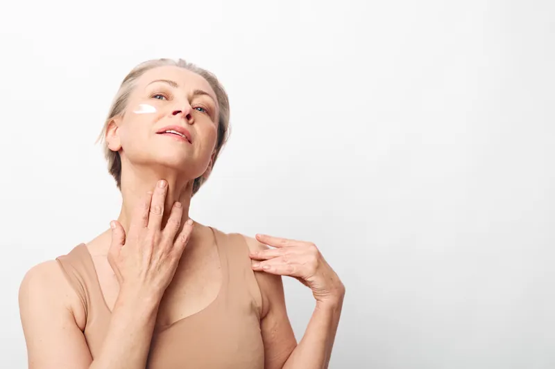 Mature woman in beige top applies white face cream on cheek and neck, demonstrating skincare routine. Bright studio background highlights natural beauty and self-care concept for aging skin.
