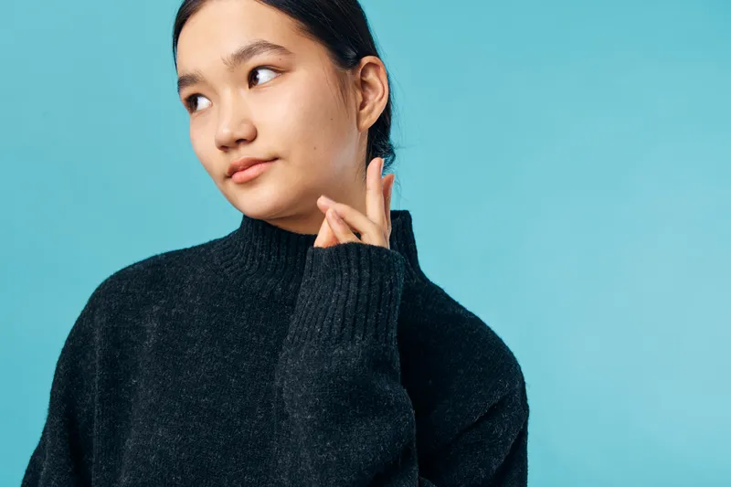 Young woman in black sweater looking sideways thoughtful expression with hand near face against blue background. Casual portrait of female model with natural light and minimal makeup.