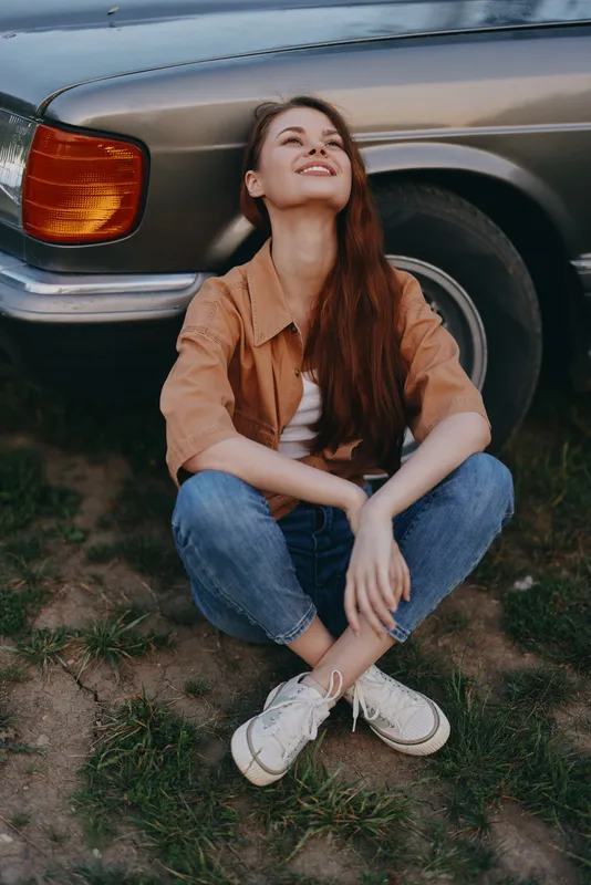 A joyful young woman with long brown hair sits by a vintage car in casual attire, radiating happiness amidst a serene outdoor setting, evoking a carefree summer vibe.