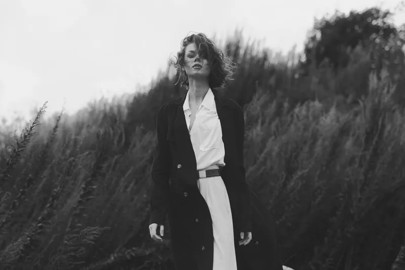 Moody black and white fashion scene with a woman in a long coat standing in tall grass, wind swept, cinematic mood, contemporary style and elegance