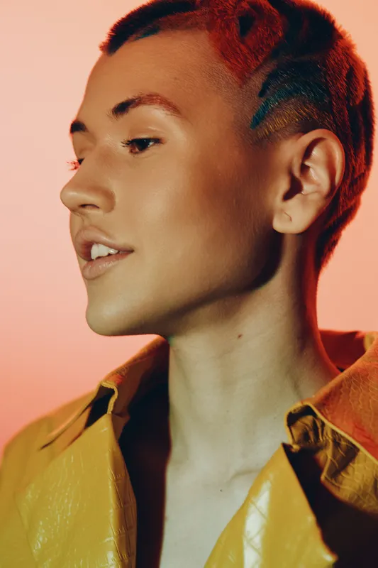 Close-up portrait of a young woman with short hair and artistic shaved patterns, wearing a vibrant yellow jacket against a warm gradient background, showcasing contemporary style.