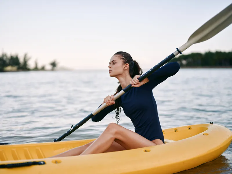 Active woman kayaking on calm water during sunset, wearing a dark blue outfit, focused and enjoying outdoor water sports.