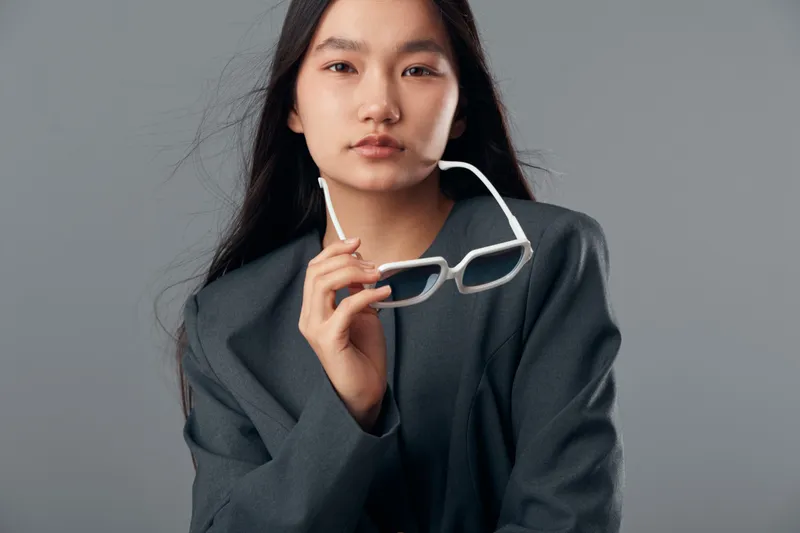 Young woman with glasses posing in oversized dark blazer against gray background. Asian female model with long hair and calm expression in casual modern outfit for portrait.