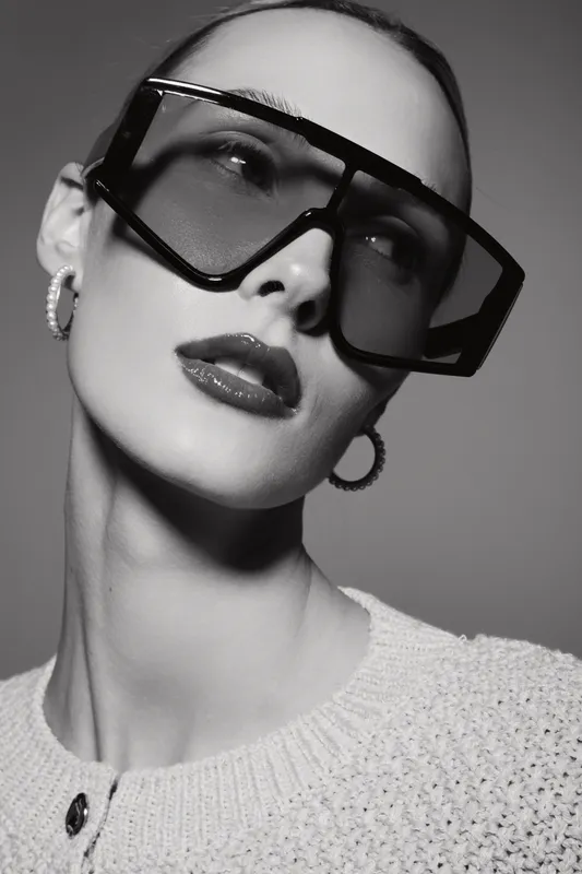 Close-up of a fair skinned Caucasian woman wearing large square sunglasses and hoop earrings. Fashion portrait with neutral expression on gray background.