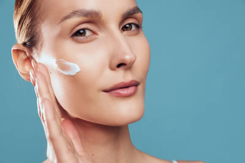 Young woman applies moisturizing cream on face with hand closeup portrait skin care concept natural beauty isolated on blue background with calm expression and soft lighting.