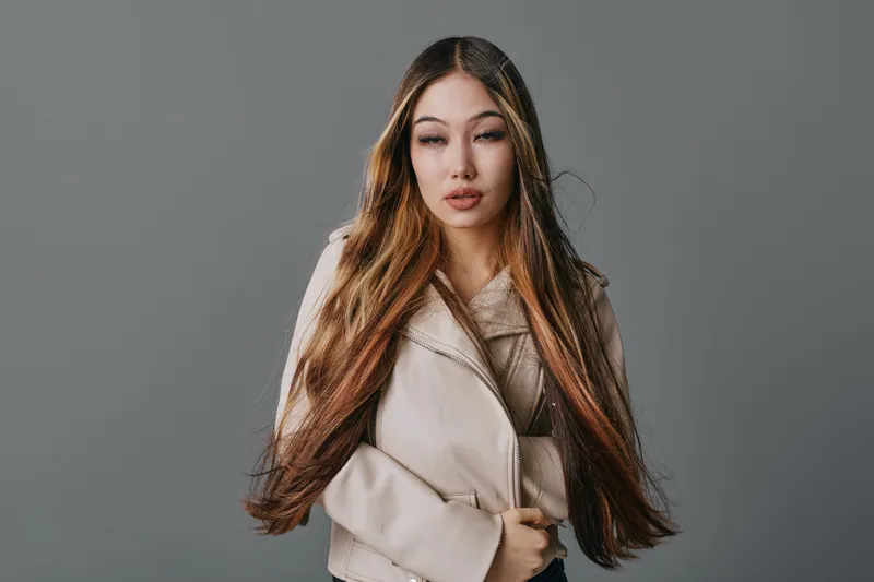 Young woman with long straight hair and beige jacket posing for portrait on gray studio background. Beauty model with fashion style and natural makeup looking at camera indoors.