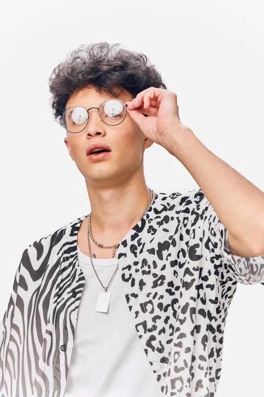 Young man wearing round glasses and a black and white animal print shirt adjusting his eyewear with a curious expression on a plain white background.