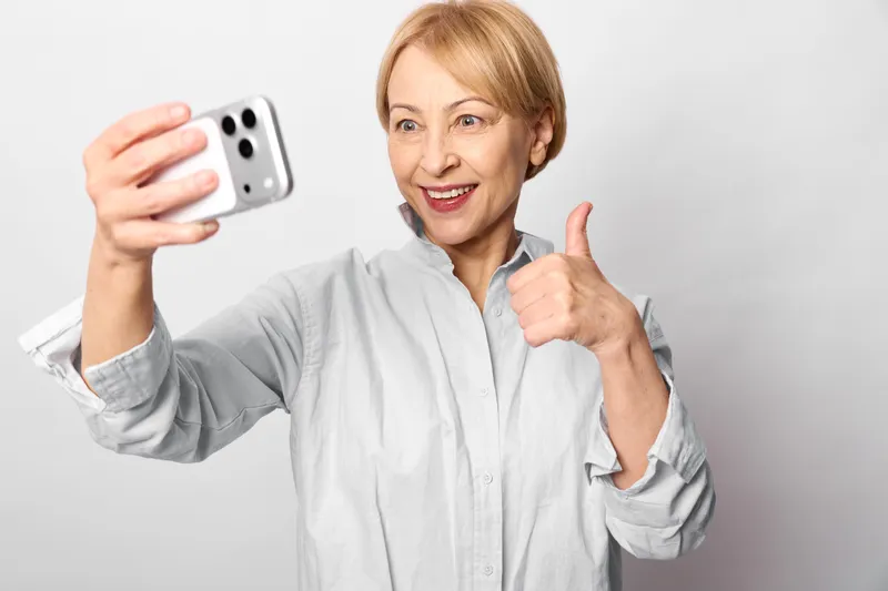 Senior woman takes a selfie with smartphone, smiling at camera, thumbs up gesture, studio portrait, white background, modern technology