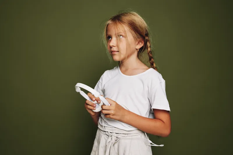 Young girl with blond hair in white t-shirt holding headphones in front of olive green background.