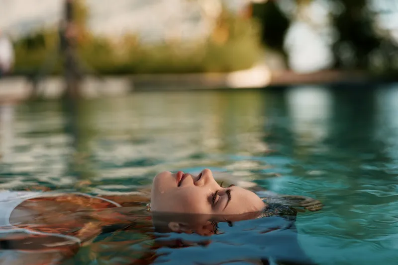 Young woman floats on her back in a clear pool with calm water around her, eyes closed, a peaceful outdoor moment captured in soft light.