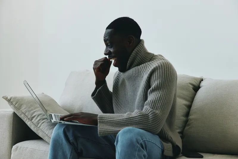Smiling young African man enjoying his time working on a laptop at home, showcasing a casual style in a cozy living room environment