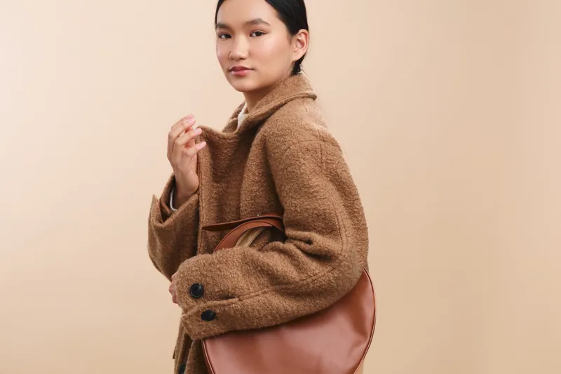Asian woman wearing a warm brown coat with a large bag poses against a soft beige backdrop, exuding confident style, poised calm, and modern casual elegance.