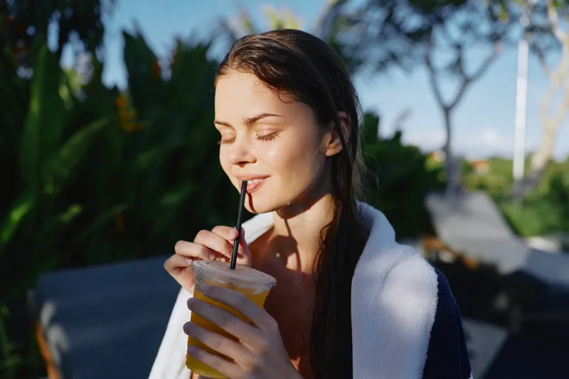 Cheerful woman outdoors, drinking a cold beverage with a straw, sunlit moment, relaxed posture, soft breeze, natural colors, lush greenery background