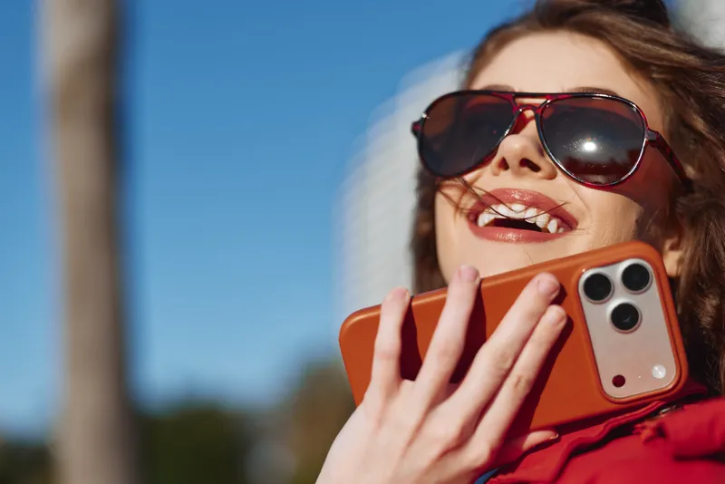 Smiling woman wearing sunglasses holds smartphone near face outdoors, enjoying sunny day with clear blue sky background, casual lifestyle and happiness concept.