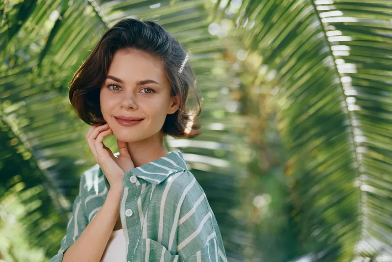 A cheerful woman outdoors among palm leaves, wearing a striped green shirt, radiating warmth and confidence.