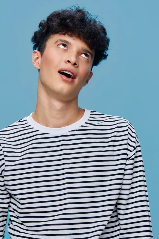 young man wearing striped shirt looking up with surprised expression on blue background, casual style and curly hair, studio portrait with bright lighting