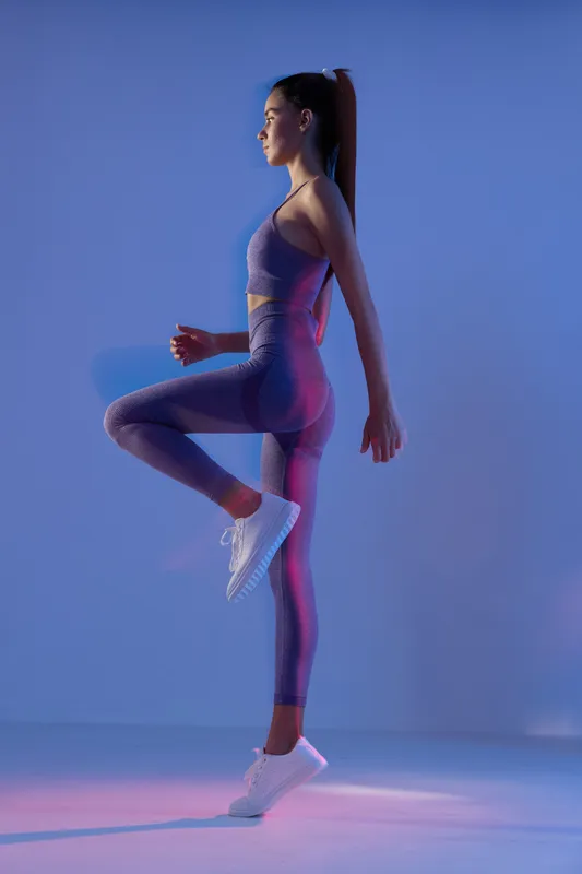 Woman sport fitness exercising indoors, wearing purple activewear and white sneakers, performing balance and strength workout in studio with ambient blue and pink lighting.